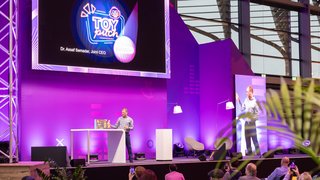 Vortrag auf lila Bühne beim Toy Business Forum, Präsentation „TOY pitch“ auf Großbildschirm.