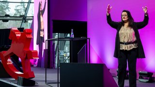 Vortragende auf Bühne mit großem Schaukelpferd-Logo und Publikum.