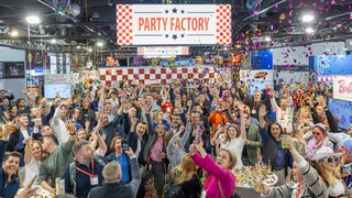 Viele Menschen feiern ausgelassen mit Konfetti unter „PARTY FACTORY“-Schild auf der Messe.