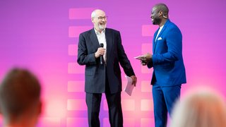 Zwei Männer im Gespräch auf Bühne, einer hält Mikrofon, lila Hintergrund.