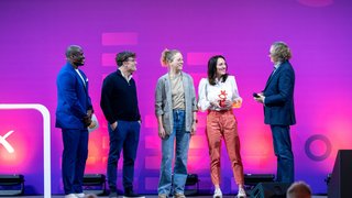 Fünf Personen stehen auf Bühne, eine hält eine Trophäe, lila Hintergrund.
