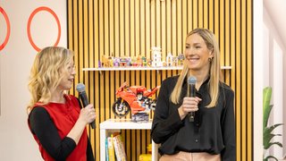 Two women with microphones speak in front of LEGO models and bookshelf at the fair.