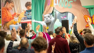 Menschen jubeln einer Tiermaskottchen-Figur auf Bühne bei Lalinos-Stand auf der Messe zu.