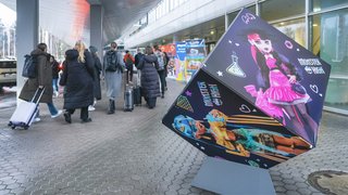 Monster High promo cube outside hall, visitors with suitcases walk past.