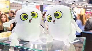 Zwei weiße Plüschfiguren mit großen gelben Augen sitzen auf einem Glastisch auf der Messe.