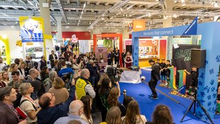 Press event at fair, many spectators and cameras at Ravensburger booth with football activity.